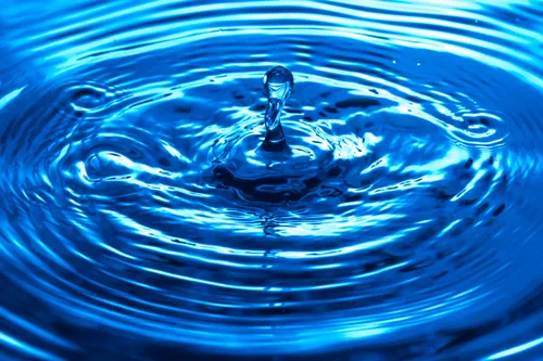 Ein Wassertropfen fällt ins blaue Wasser und erzeugt Wellen und Ringe. Das Wasser ist klar und blau.