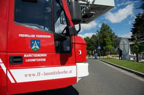 Die Vorderseite eines roten Feuerwehrwagens mit 'WILLIGE FEUERWEHR' und 'MARKTGEMEINDE LASSNITZHOEHE' Text. Ein Logo mit Kreuz und Baum ist auf dem Wagen. Im Hintergrund sind Menschen, Bänke und Bäume.