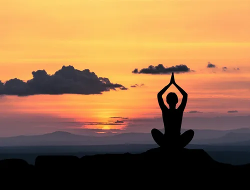 Eine Silhouette einer Person, die bei Sonnenuntergang Yoga in Lotusposition macht. Die Person sitzt auf einem Berggipfel.