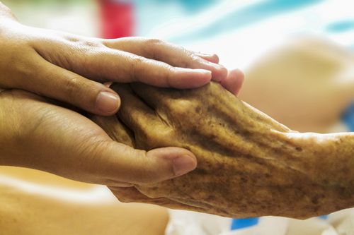 Ein Nahaufnahme einer Hand, die eine ältere Person tröstet, zeigt Hauttextur und Falten. Die Szene ist drinnen, mit einem unscharfen Hintergrund.