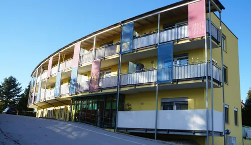 Ein gelbes Mehrfamilienhaus mit mehreren Balkonen, jeder mit einem blauen Vorhang, vor einem klaren blauen Himmel.