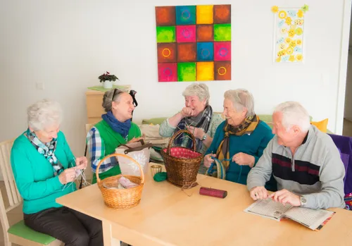 Eine Gruppe älterer Frauen und Männer sitzt um einen Tisch herum, strickt, mit Körben und einem farbenfrohen Gemälde an der Wand.