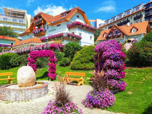 Eine malerische Dorfszene mit einem charmanten weißen Haus, 'Enifenheim' darüber geschrieben. Bunte Blumen schmücken die Balkone. Eine Holzbank ist zwischen üppiger Grünanlage eingebettet.