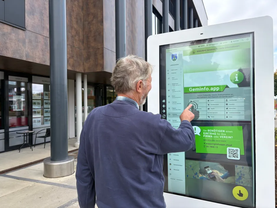 Ein älterer Mann benutzt einen Touchscreen-Terminal im Freien. Der Bildschirm zeigt Informationen über eine lokale App, einschließlich eines QR-Codes für die Registrierung.