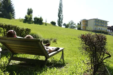 Eine Person ruht sich auf einer Bank in einem Grasfeld aus, mit einem Gebäude im Hintergrund.