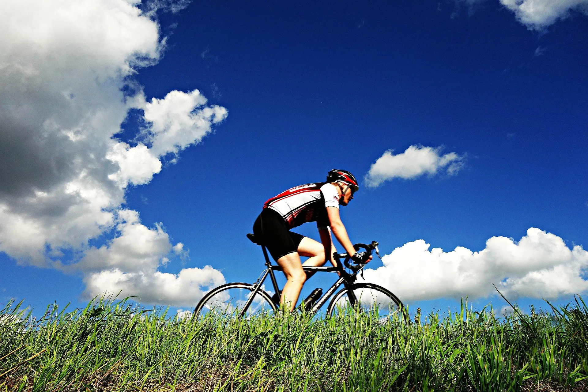 Ein Radfahrer fährt auf einem Rasenfeld einen Hügel hinauf, während er einen Fahrradhelm und eine Radsportausrüstung trägt. Der Himmel ist blau mit einigen Wolken.