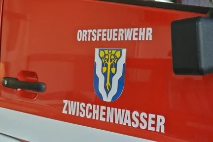 Ein rotes Fahrzeug, wahrscheinlich ein Feuerwehrauto, hat ein Logo mit einem Baum und der Aufschrift 'ORTSFEUERWEHR' darüber und dem Wort 'ZWISCHENWASSER' darunter.