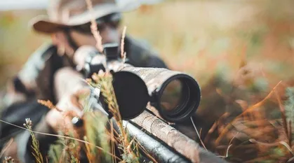 Eine Person in Tarnkleidung und einem Hut hält ein Gewehr mit Zielfernrohr auf einem Feld.