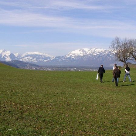 Vier Personen laufen über einen grasbewachsenen Hügel mit verschneiten Bergen im Hintergrund. Sie sind für kühles Wetter gekleidet.