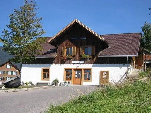 Ein traditionelles Holzhaus mit braunem Dach und weißen Wänden, auf einem Hang mit Grün und einem Baum gelegen. Kreuze sind an den Fenstern zu sehen.
