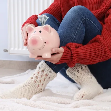 Eine Frau sitzt auf einem Bett, trägt einen roten Pullover und Jeans und hält eine Sparschwein-Bank mit Geld, das herausragt.