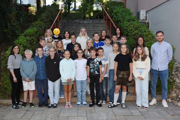 Eine Gruppe von Kindern, Lehrern und einem Erwachsenen Mann posiert für ein Foto vor einem Gebäude, mit einer Treppe und Grünpflanzen im Hintergrund.