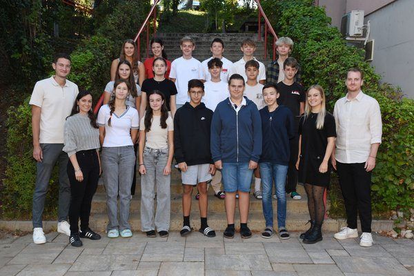 Eine Gruppe von Schülern und Lehrern posiert für ein Foto an einem sonnigen Tag, mit Treppen und Grünpflanzen im Hintergrund.