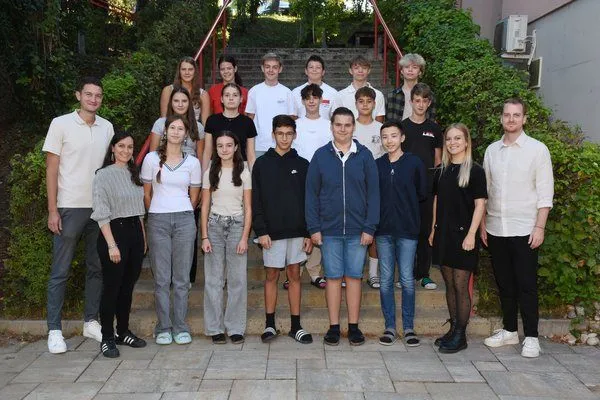 Eine Gruppe von Schülern und Lehrern posiert für ein Foto an einem sonnigen Tag, mit Treppen und Grünpflanzen im Hintergrund.