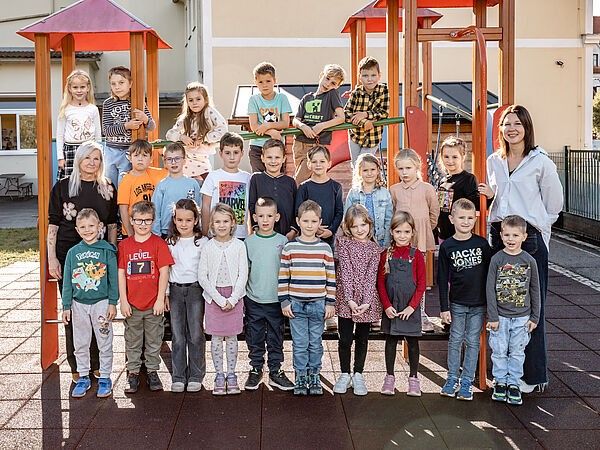 Eine Gruppe von Kindern und zwei Frauen stehen vor einem Spielplatz. Die Kinder tragen Freizeitkleidung. Die Frauen lächeln und stehen neben den Kindern.