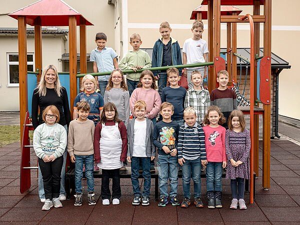 Eine Gruppe junger Schüler und eine Frau stehen vor einem Spielplatz mit einem Gebäude im Hintergrund.