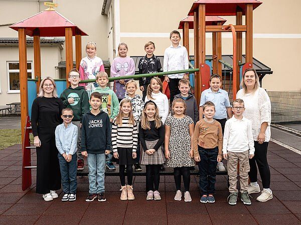 Eine Gruppe von Kindern und zwei Erwachsenen steht vor einem Spielplatz mit verschiedenen Spielgeräten im Hintergrund.