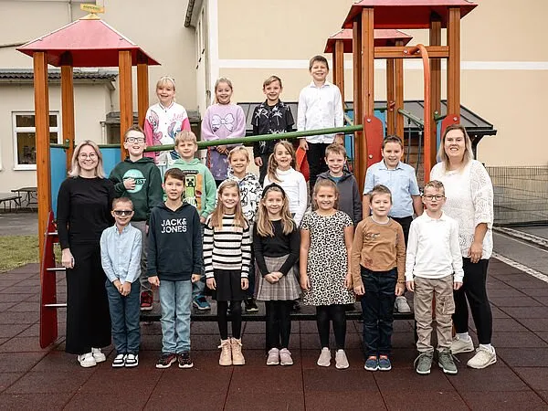 Eine Gruppe von Kindern und zwei Erwachsenen steht vor einem Spielplatz mit verschiedenen Spielgeräten im Hintergrund.