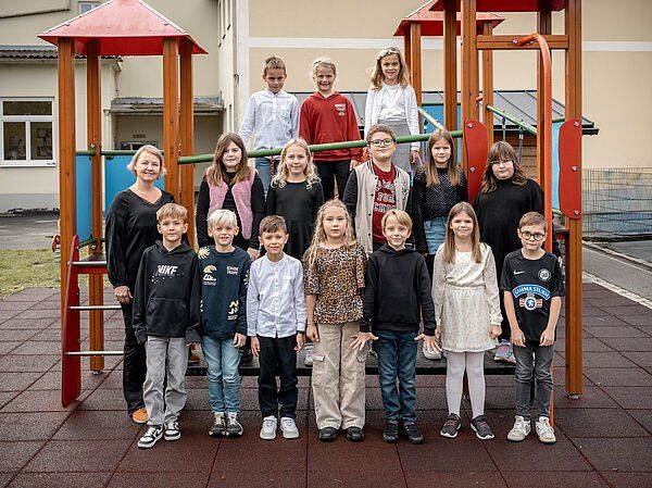 Eine Gruppe von Kindern und zwei Frauen posiert für ein Foto vor einem Spielplatz mit einem Gebäude im Hintergrund.