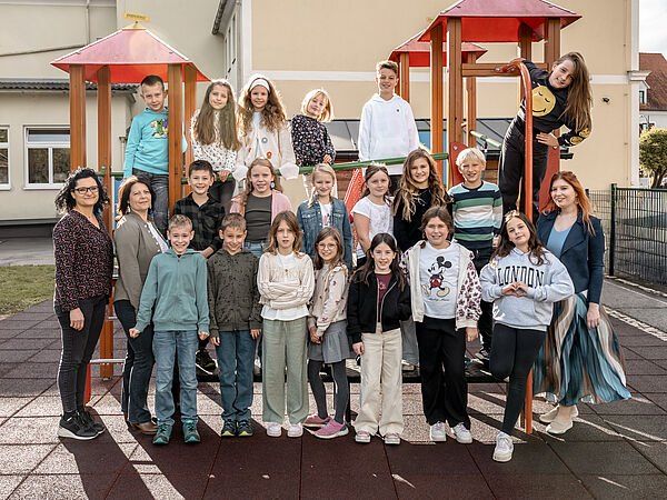 Eine Gruppe von Kindern und zwei Erwachsenen steht in einem Spielplatz mit bunten Spielstrukturen im Hintergrund. Die Kinder lächeln und posieren für die Kamera.