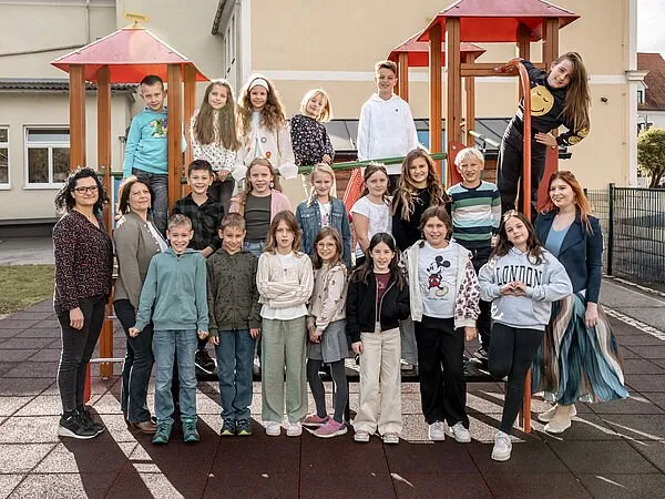 Eine Gruppe von Kindern und zwei Erwachsenen steht in einem Spielplatz mit bunten Spielstrukturen im Hintergrund. Die Kinder lächeln und posieren für die Kamera.