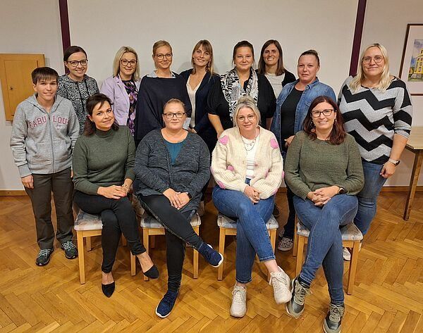 Eine Gruppe von Frauen und einem Mann posiert für ein Foto in einem Raum, alle in lässiger Kleidung. Sie sitzen auf Holzstühlen und stehen vor einer weißen Wand.