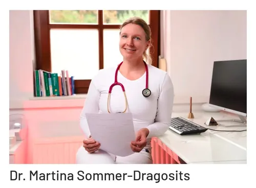Eine Frau in weißer Uniform und mit Stethoskop, lächelnd, hält ein Papier vor einem Computer. Hinter ihr sind Bücher und ein Fenster mit Blick auf Bäume zu sehen.