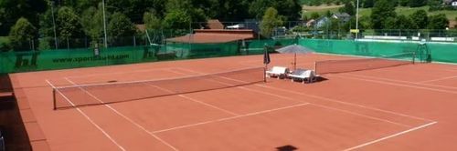 Eine Vogelperspektive eines leeren Tennisplatzes mit rotem Lehmboden und weißen Linien. Zwei Liegestühle stehen in der Nähe des Zauns, und ein Regenschirm ist an der Seite.