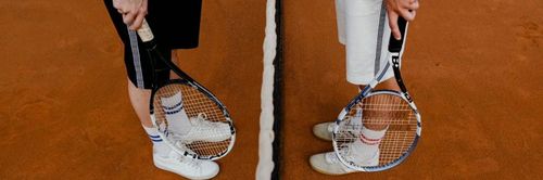 Die Beine eines Tennisspielers sind in weißen Schuhen, Socken und einem Schläger zu sehen. Der Hintergrund ist ein Tennisplatz.