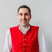 Ein Mann in einem roten Weste mit weißen Knöpfen und einem weißen Langarmhemd lächelt vor einer weißen Wand.