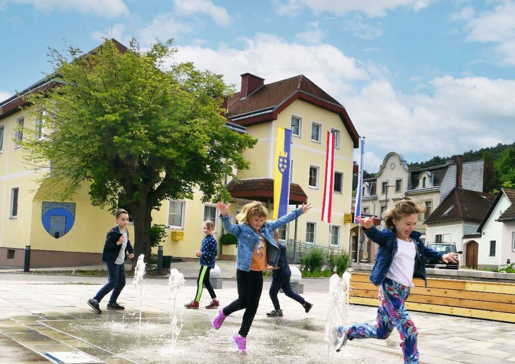  Abschnitt Bild: Kinder spielen und springen in einem Brunnen vor einem gelben Gebäude mit Fahnen. Bäume und ein Auto sind im Hintergrund zu sehen.