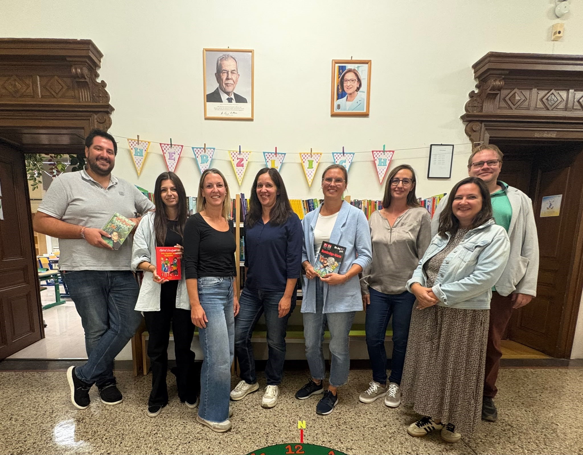 Eine Gruppe von Menschen in einer Bibliothek, lächelnd und Bücher haltend, mit Wimpelketten und gerahmten Porträts im Hintergrund.