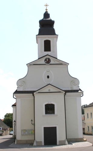 Eine kleine weiße Kirche mit einem schwarzen Turm und einer Uhr. Vorne befindet sich eine schwarze Tür.