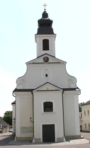 Eine kleine weiße Kirche mit einem schwarzen Turm und einer Uhr. Vorne befindet sich eine schwarze Tür.