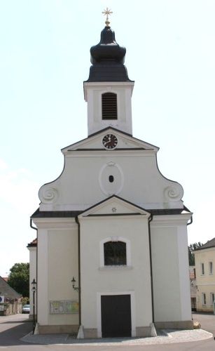Eine kleine weiße Kirche mit einem schwarzen Turm und einer Uhr. Vorne befindet sich eine schwarze Tür.