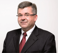 Ein Mann in Anzug und Krawatte lächelt für ein Foto. Er trägt eine Brille und hat graues Haar.
