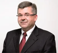 Ein Mann in Anzug und Krawatte lächelt für ein Foto. Er trägt eine Brille und hat graues Haar.