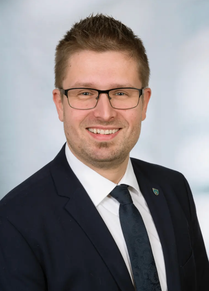 Ein Mann in einem dunklen Anzug und mit Brille lächelt in die Kamera. Er trägt ein weißes Hemd und eine dunkle Krawatte. Auf seinem linken Revers befindet sich ein Logo.