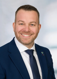 Ein Mann in Anzug und Krawatte, lächelnd, mit einem hellblauen Himmel im Hintergrund.