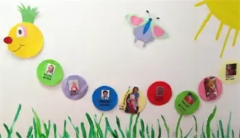 Eine weiße Wand zeigt einen Raupe mit bunten Punkten, die jeweils ein Foto enthalten. Die Raupe besteht aus Papier und beinhaltet einen Schmetterling und eine Sonne. Unten sind grüne Grashalme.