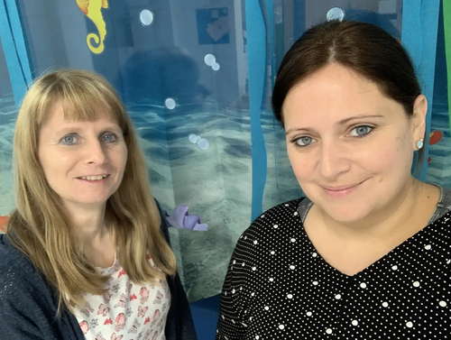 Zwei Frauen stehen eng beieinander, lächeln und posieren für ein Foto. Die Frau auf der linken Seite hat blonde Haare. Sie stehen vor einer blauen Wand mit einem Meeresdesign.