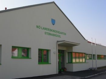 Ein weißes Gebäude mit grünen Fenstern und einem blauen Schild über dem Eingang, das 'NO LANDESKINDERGARTEN STEINABRUCKL' steht.