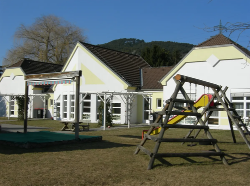 Ein Spielplatz mit Rutsche und Schaukel steht vor einem Haus mit braunem Dach. Das Haus hat weisse Wände und gelbe Akzente.