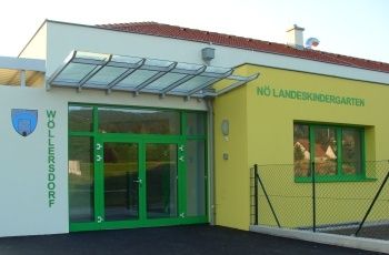 Ein modernes Gebäude mit gelber Außenseite und grünen Glastüren. Es hat ein Wappenlogo auf der linken Seite und ein rotes Dach. Das Gebäude ist als 'No Landeskindergarten' beschriftet. Rechts befindet sich ein Metallzaun.