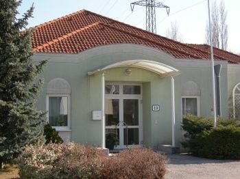 Ein weißes Gebäude mit einem roten Ziegeldach und Glastüren und -fenstern. Die Nummer 18 befindet sich über der Tür. Über dem Eingang befindet sich ein kleiner Vorbau. Vor dem Gebäude stehen Büsche und Bäume.