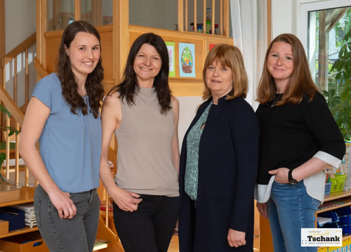 Vier Frauen stehen in einem Raum mit einer Treppe und Regalen. Sie tragen alle Freizeitkleidung. Die Frau auf der linken Seite hat lange Haare und trägt ein blaues Shirt und Jeans. Die Frau neben ihr trägt ein graues Oberteil und schwarze Hose. Die Frau auf der rechten Seite trägt ein schwarzes Oberteil und Jeans. Die Frau in der Mitte trägt einen schwarzen Mantel.