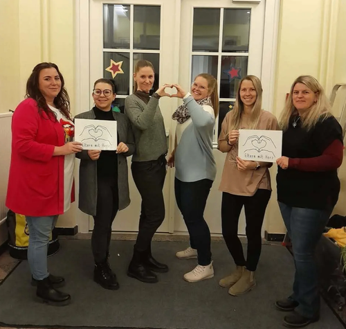 Fünf Frauen stehen vor einer weißen Tür mit roten Sternen. Jede hält ein herzförmiges Schild mit den Worten 'Eltern mit Herz' in der Hand.