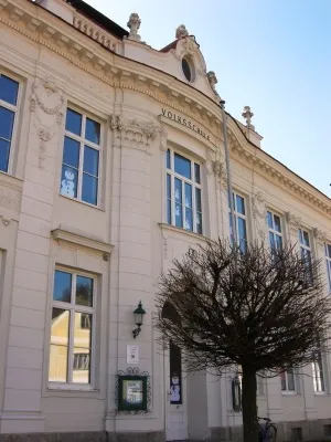 Ein großes weißes Gebäude mit vielen Fenstern und kunstvollen Verzierungen über dem Eingang, einem Baum davor und einer Lampe neben der Tür.