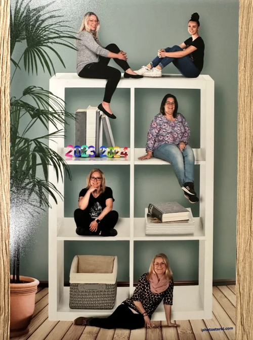Vier Frauen posieren auf einem Bücherregal, eine sitzt auf dem unteren Regal, zwei auf den mittleren Regalen und eine auf der oberen. Zwei Regale haben Bücher. Die Jahre 2023 und 2024 sind auf dem mittleren Regal zu sehen.