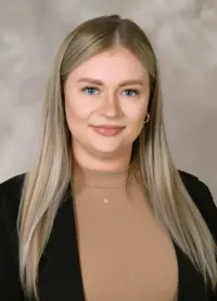 Eine Frau mit langen blonden Haaren und einem Nasenring posiert für ein Foto in einem schwarzen Blazer und einem braunen Rollkragenpullover.